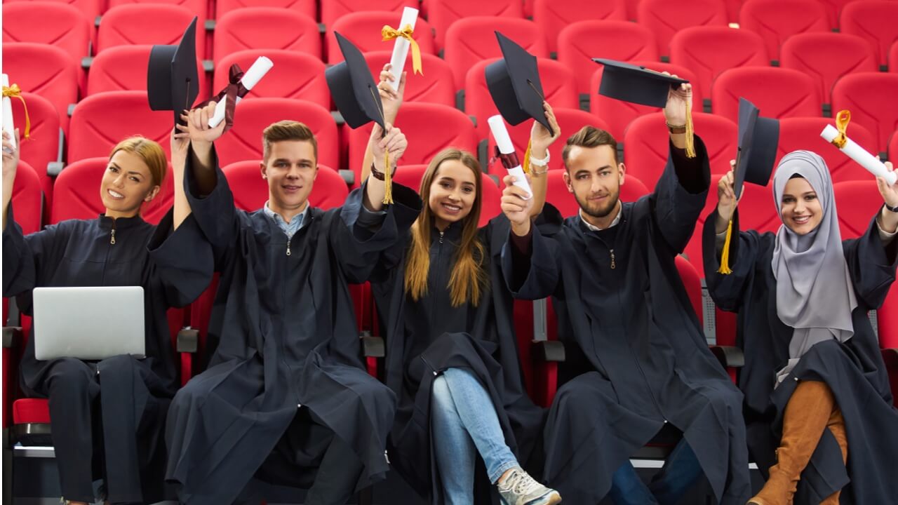 college grads sitting holding caps up with red seats behind