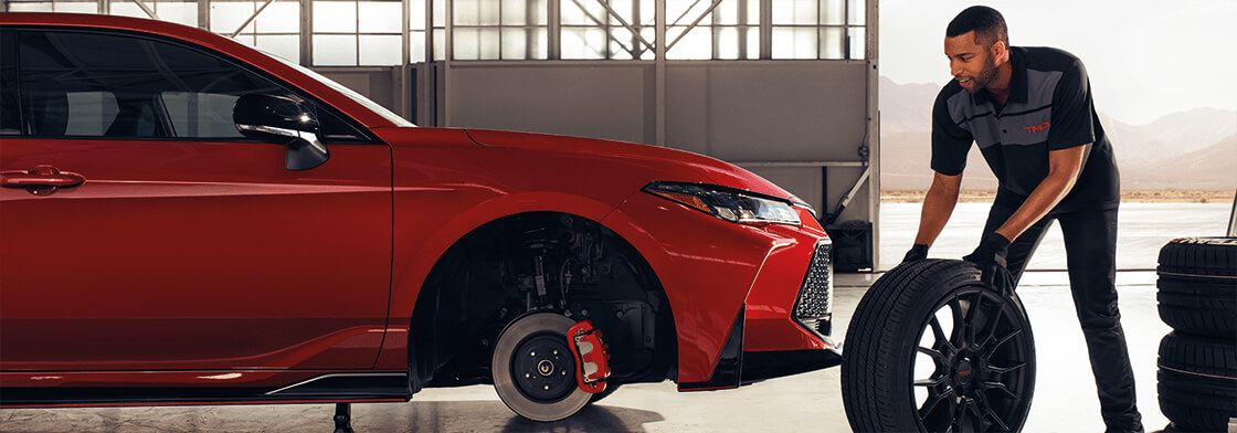 A mechanic rolls a wheel toward a red Toyota vehicle in an open garage. 