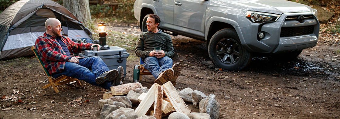 Two men sit by a campfire in the forest, surrounded by a camping tent to the left and a 4Runner to the right. 