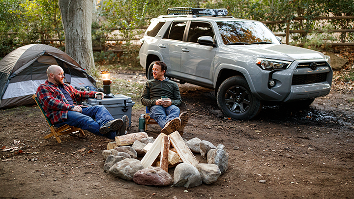 Two men sit by a campfire in the forest, surrounded by a camping tent to the left and a 4Runner to the right. 