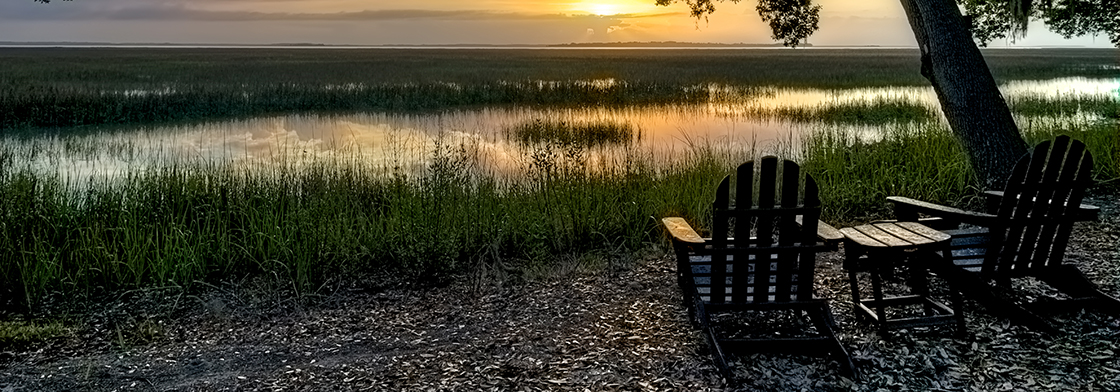 Two wooden lawn chairs sit in front of a marshland at sunset. 