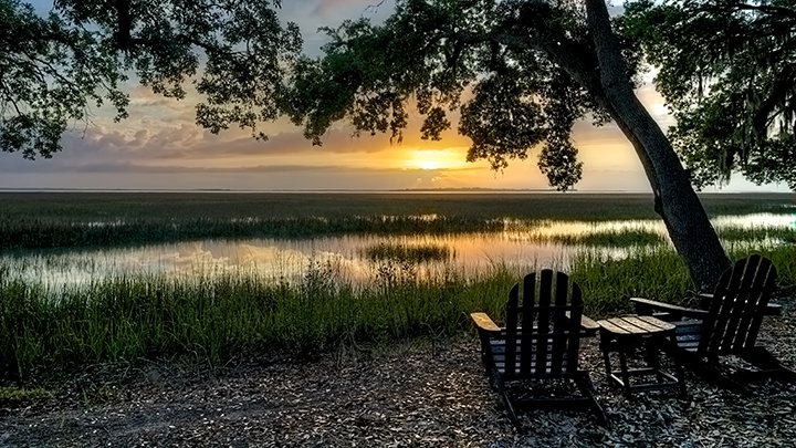 Two wooden lawn chairs sit in front of a marshland at sunset. 