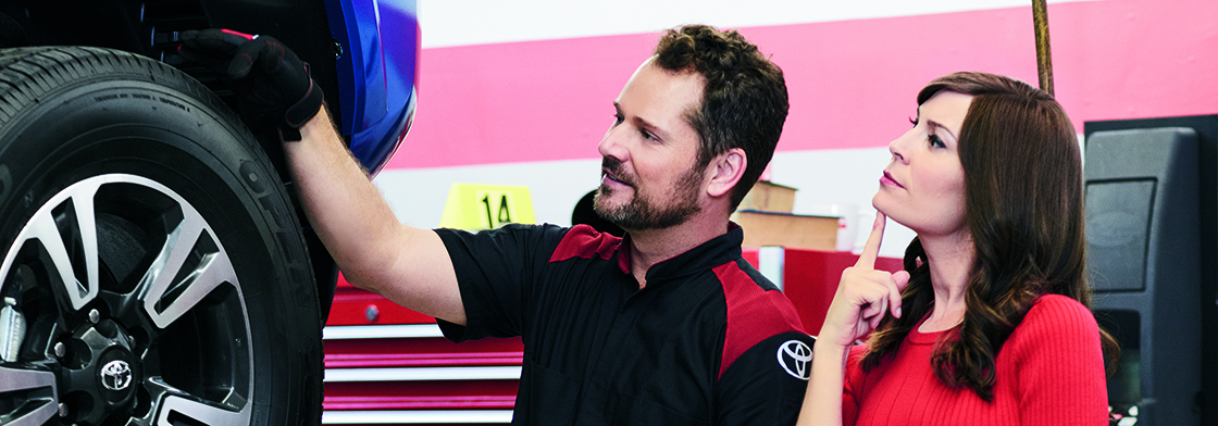 A man rests his hand on the tire of a car while a woman stands beside him, looking contemplative with a finger to her chin. 